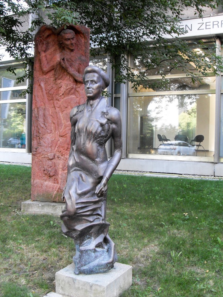 Skulpturen am Franz-Mehring Platz: Rosa Luxemburg von Rolf Biebl, dahinter eins von zwei Terrakotta-Reliefs zu Ehren von Mathilde Jacob und Karl Liebknecht von Ingeborg Hunzinger